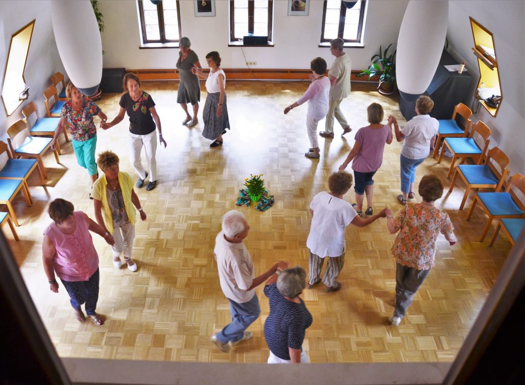 Eine Gruppe Menschen tanzt in einem großen hellen Raum im Kreis