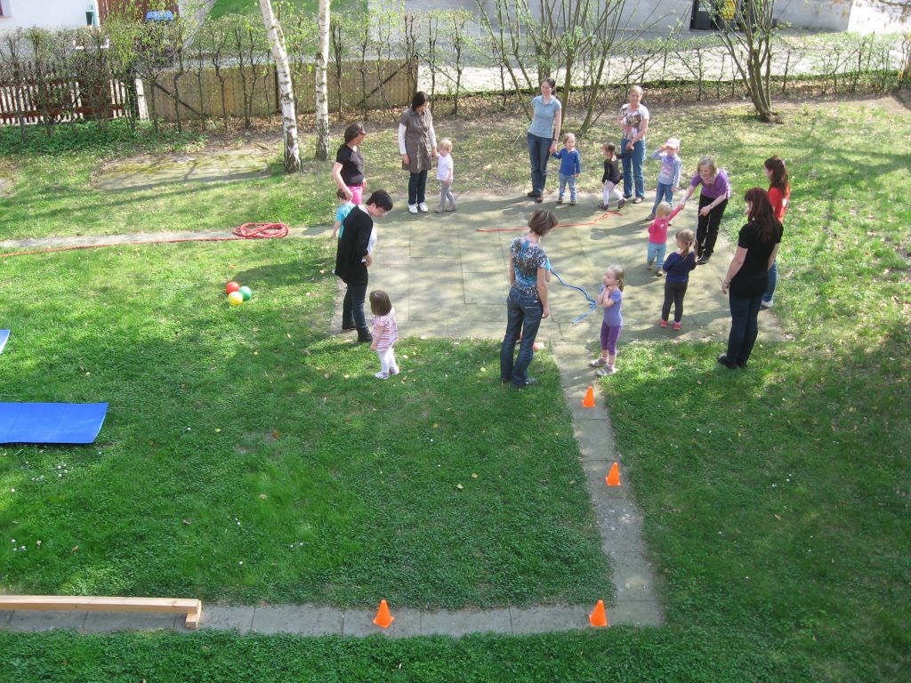 Kursbild Kinder-Sport - jeweils eine erwachsene Person steht mit einem Kind zusammen. Die ganze Gruppe steht im Kreis im Freien auf einer Wiese. Es sind verschieden kleine Sportgeräte zu sehen. 